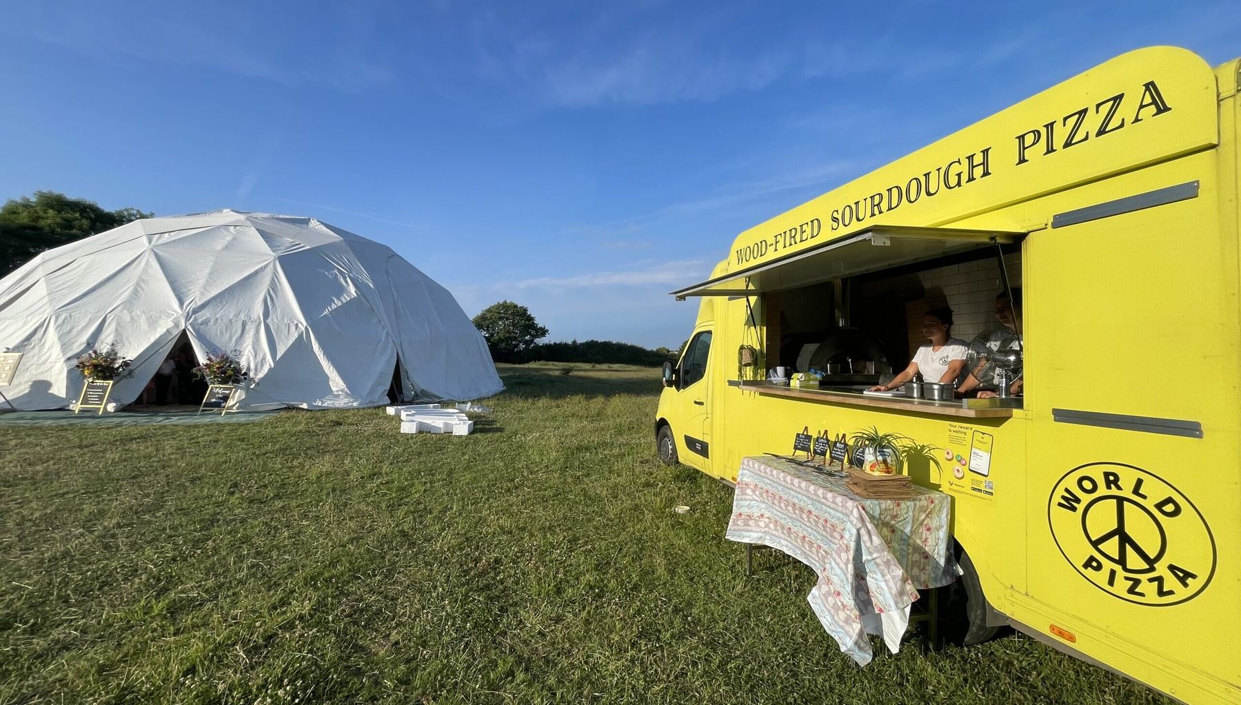 Margot, the yellow World Pizza van, serving at an outdoor wedding beside a white dome marquee.
