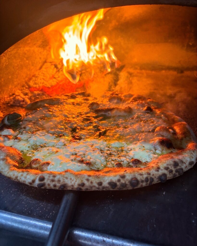 A pizza blistering in the woodfired oven with flames rolling across the roof of the oven.